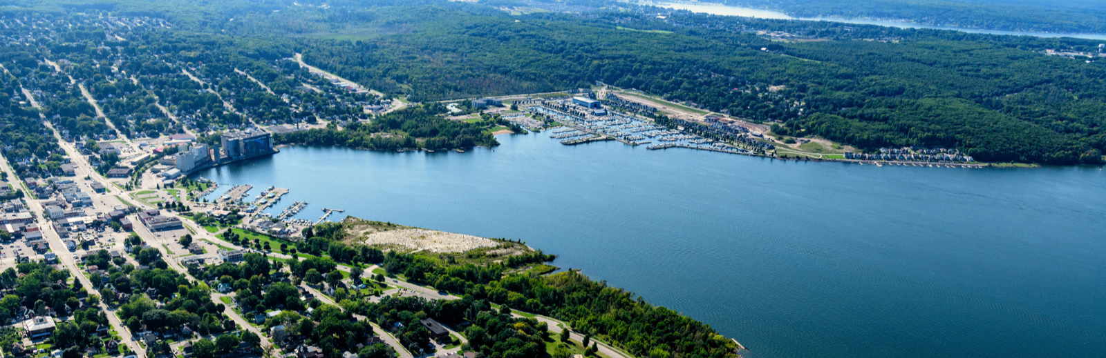 An aerial view of a waterfront town with a large bay surrounded by marinas, docks, and boats. Residential streets and buildings line the shoreline on the left, while dense green forest stretches across the background. The water is calm and deep blue under clear skies.