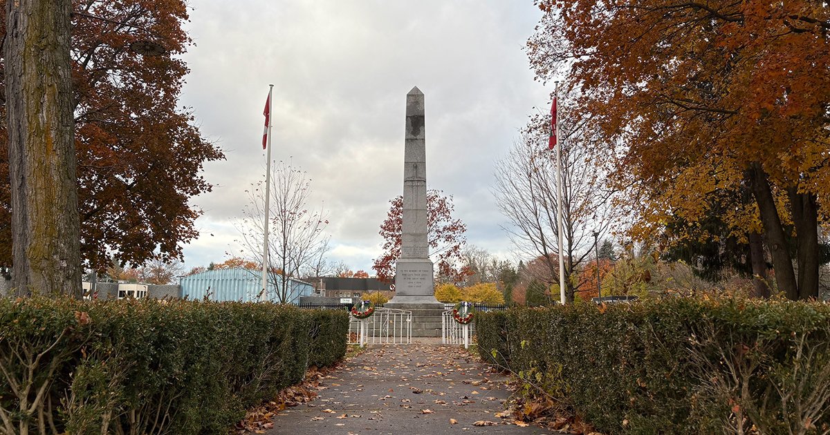 A tall stone cenotaph monument stands at the center of a paved pathway, flanked by trimmed hedges and two Canadian flags on poles. Wreaths are placed at the base of the monument, and autumn trees with orange and red leaves surround the scene. The sky is overcast with light gray clouds.