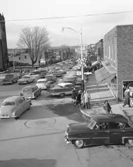 A historic black-and-white photograph of downtown Midland showing a busy main street filled with 1950s-era cars, storefronts, and pedestrians. Vehicles line both sides of the street, while vintage business signs and brick buildings stretch into the distance toward the waterfront.