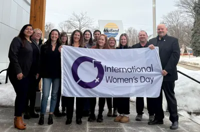 A group of people standing outside a building in winter, holding a white flag that reads “International Women’s Day” with a purple circular symbol. Snow surrounds the area, and the Midland logo is visible in the background.