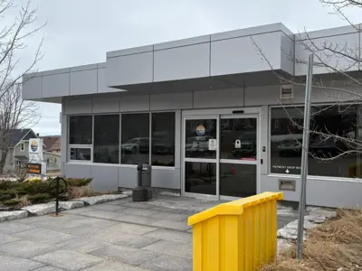The entrance to the Midland Municipal Office on an overcast day, showing glass double doors, large windows, a paved walkway, and a bright yellow salt storage container in the foreground.
