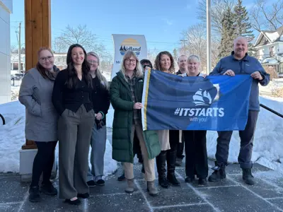 A group of people standing outside a building in winter, holding a blue flag that reads “#ITSTARTS with you!” Snow covers the ground around them, and a Midland banner is displayed in the background.