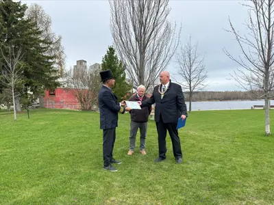 Three people stand on a grassy waterfront park during an outdoor presentation, with one person handing a certificate to another, who's wearing a top hat. Trees, a historic red building, and calm water are visible in the background under an overcast sky.