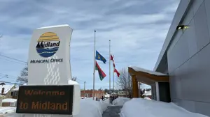 The Midland Municipal Office entrance in winter, with three flags flying at half‑mast beside the walkway. A large roadside sign reads “Welcome to Midland,” and deep snow lines the path under a partly cloudy sky.