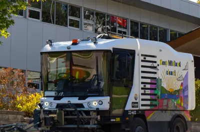 A white street sweeper vehicle moving near a modern building, featuring a colourful mural and “Keep Midland Clean” text on the side.