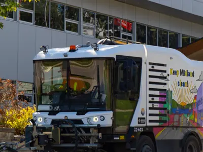 A white street sweeper vehicle moving near a modern building, featuring a colourful mural and “Keep Midland Clean” text on the side.
