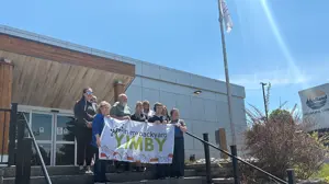 A group of people stand on the steps outside a municipal building holding a banner that reads “Yes in my backyard – YIMBY.” A flagpole and Midland signage are visible nearby under a bright blue sky.