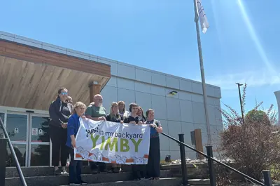 A group of people stand on the steps outside a municipal building holding a banner that reads “Yes in my backyard – YIMBY.” A flagpole and Midland signage are visible nearby under a bright blue sky.