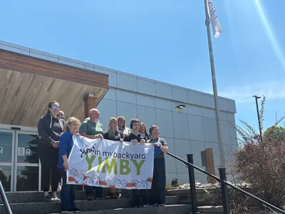 A group of people stand on the steps outside a municipal building holding a banner that reads “Yes in my backyard – YIMBY.” A flagpole and Midland signage are visible nearby under a bright blue sky.