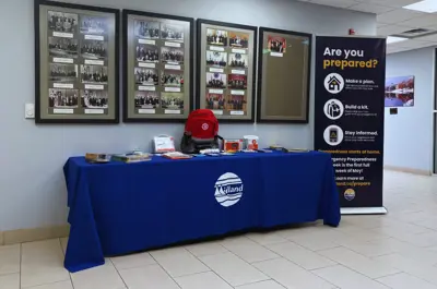 An indoor emergency preparedness display featuring a table with a blue Midland‑branded tablecloth, brochures, a red emergency kit backpack, and preparedness supplies. Behind the table are framed historical photos on the wall and a standing banner that reads “Are you prepared?” with tips for making a plan, building a kit, and staying informed.