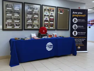 An indoor emergency preparedness display featuring a table with a blue Midland‑branded tablecloth, brochures, a red emergency kit backpack, and preparedness supplies. Behind the table are framed historical photos on the wall and a standing banner that reads “Are you prepared?” with tips for making a plan, building a kit, and staying informed.