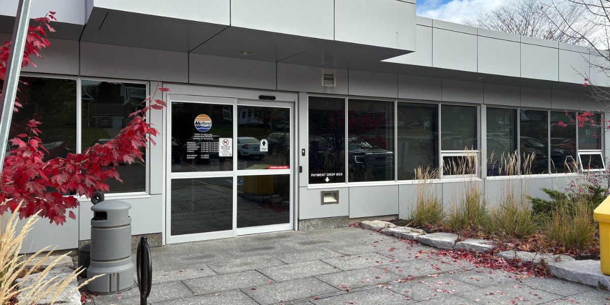 A modern, single-story building with a flat gray facade and large windows along the front. The entrance features glass double doors with signage, and a gray trash bin is positioned near the doorway. Red autumn leaves and ornamental grasses frame the walkway, which is paved with stone tiles. A bright yellow bin is visible on the right side, and the sky above is partly cloudy.